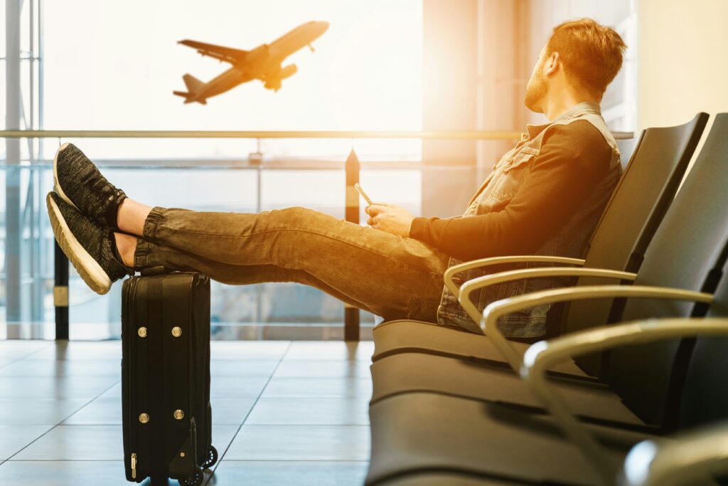 An image showing a man sitting in an airport waiting room with his luggage and an aeroplane taking off in the background