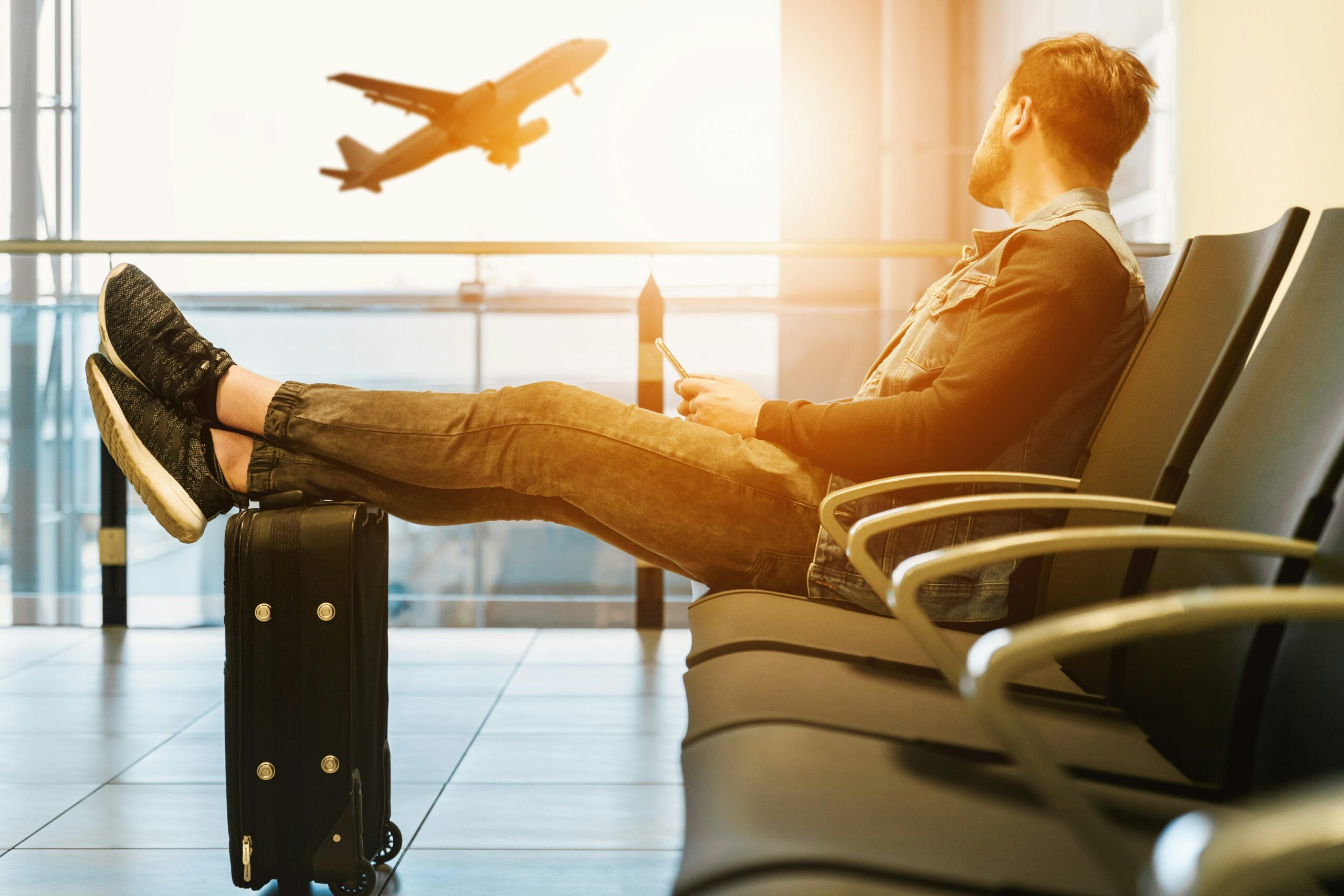 An image showing a man sitting in an airport waiting room with his luggage and an aeroplane taking off in the background